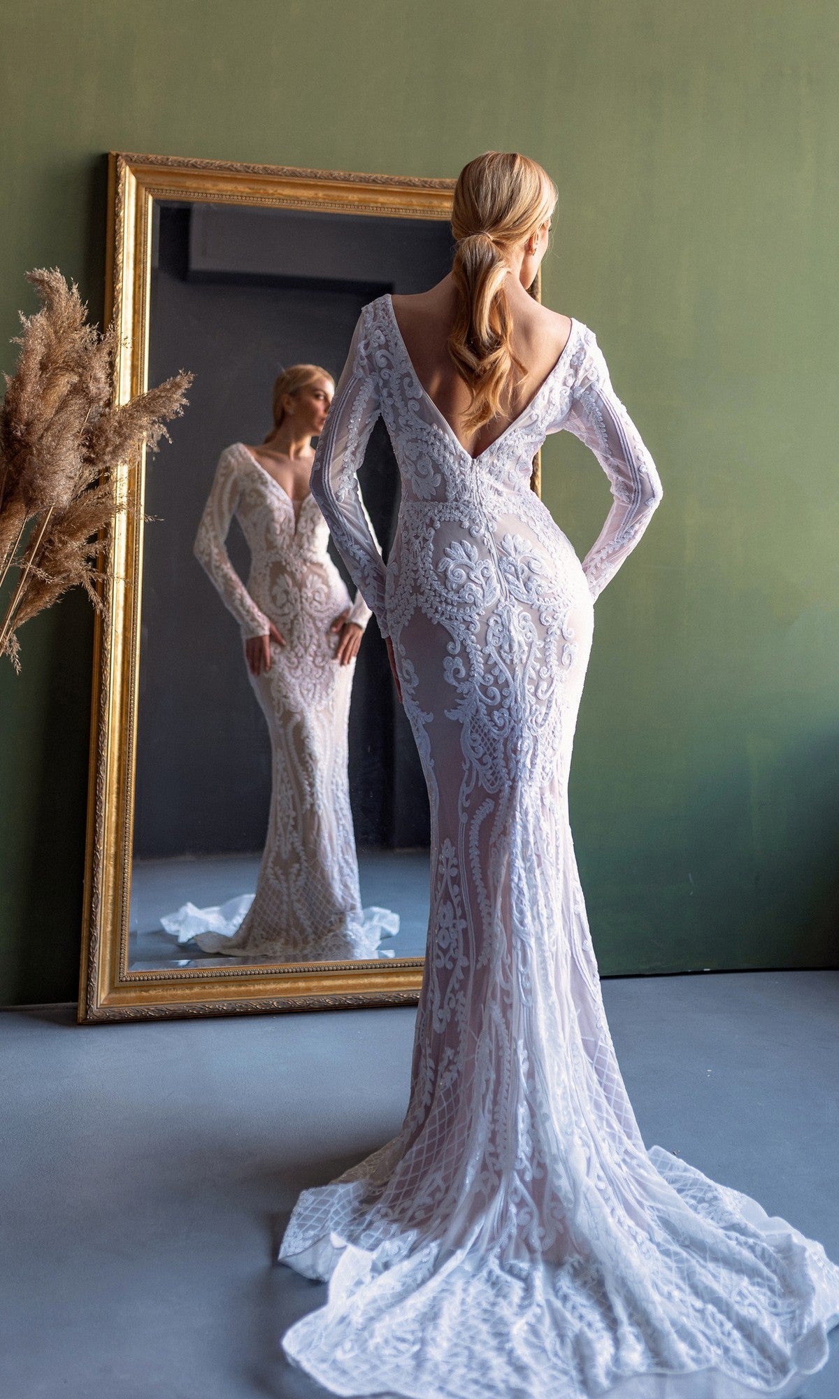 A woman wearing the Long Sleeve White Bridal Gown: Tina Holly BB021 stands facing a mirror, her reflection visible, in a room with a green wall and dried plants.